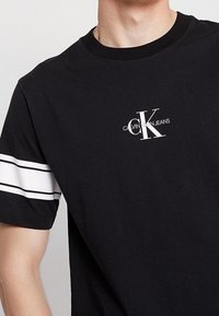 Camiseta negra con rayas blancas en las mangas, con un logo blanco de Calvin Klein Jeans destacado en el pecho. Material de algodón, diseño de cuello redondo.