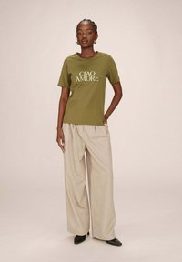 T-shirt vert olive avec le texte 'CIAO AMORE', associée à un pantalon large beige et des ballerines noires à bout pointu. Tissu lisse, coupe décontractée.