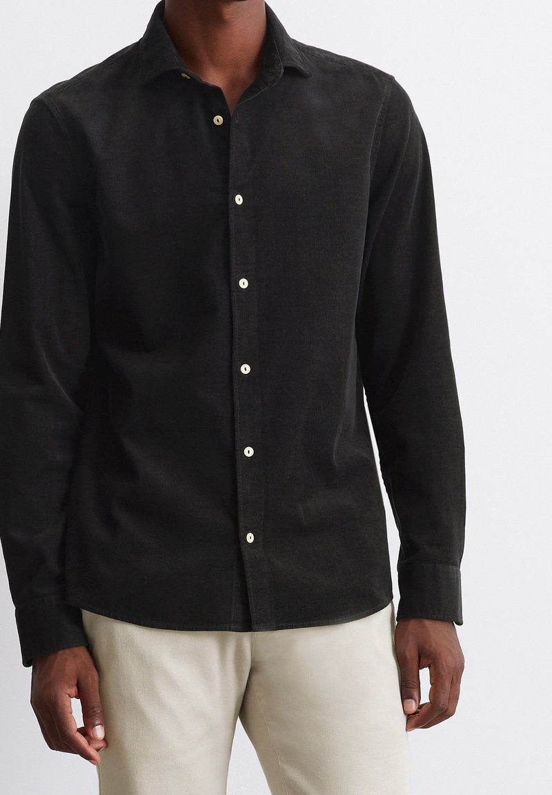 Homme portant une chemise noir en velours côtelé ajustée avec des boutons blancs et un pantalon beige clair sur un fond uni.
