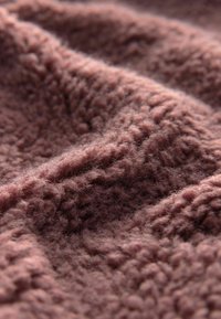 Zbliżenie na miękką, teksturowaną dzianinę w kolorze mauve, z widocznymi włóknami i delikatnymi fałdami, które tworzą głębię i cienie.