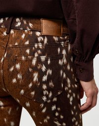 Jeans din denim cu un model maro și stropi albi, având o suprafață texturată și un patch din piele cu logo-ul Wrangler pe talie.