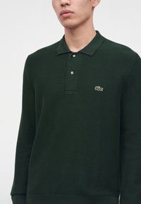 Homme portant un polo à manches longues vert foncé avec deux boutons et un petit logo crocodile sur la poitrine.