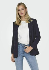 Jūras spēku žakete ar piegriezumu, ar vērto atlokiem un divām priekšējām kabatām, kombinēta ar baltu T-kreklu un gaiši ziliem džinsiem.