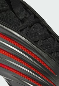Sort træningssko med tekstureret tekstiloverdel, elastiske snørebånd og blank sort sål med røde mesh-detaljer og bølgelignende design.