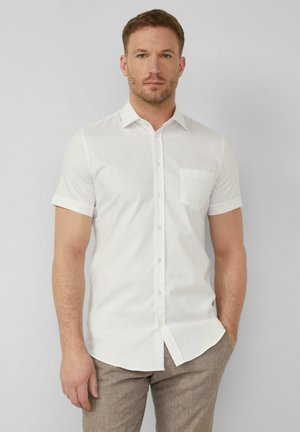 Mężczyzna w białej koszuli z krótkim rękawem i kieszenią, stojący na tle jednolitego jasnoszarego tła.