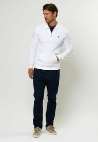 Biela mikina na zips s polovičným golierom, s klokanou vreckom. Spárovaná s tmavomodrými džínsami a svetlými teniskami.