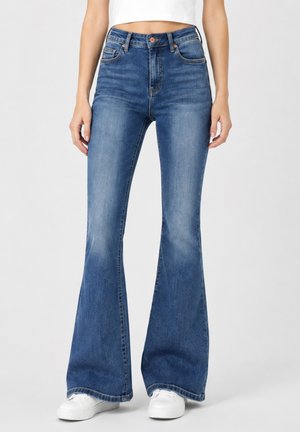 Vrouw draagt hoge blauwe flare jeans en witte sneakers, staand tegen een effen lichte achtergrond.