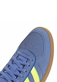 Zapatilla de ante azul con suela de goma, con rayas amarillas, un cuello acolchado y perforaciones en la punta para transpirabilidad.