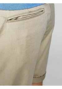 Beiget shortsit, joissa on yksinkertainen muotoilu, kaartuva takatasku ja käännetty helma. Kangas näyttää sileältä ja siinä on vähän tekstuuridetaljeja.