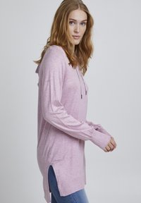 Hoodie rose clair à manches longues avec une fente sur le côté, des détails de cordon de serrage et des poignets côtelés. Le matériau semble doux avec une finition texturée.