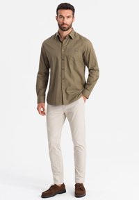 Camisa verde oliva de botones, con mangas largas y un bolsillo en el pecho, combinada con pantalones beige claros y zapatos de ante marrones.