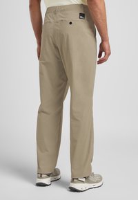 Pantaloni beige leggeri con una vestibilità comoda, dotati di una tasca posteriore, etichetta con logo e una superficie liscia, abbinati a sneakers grigie.