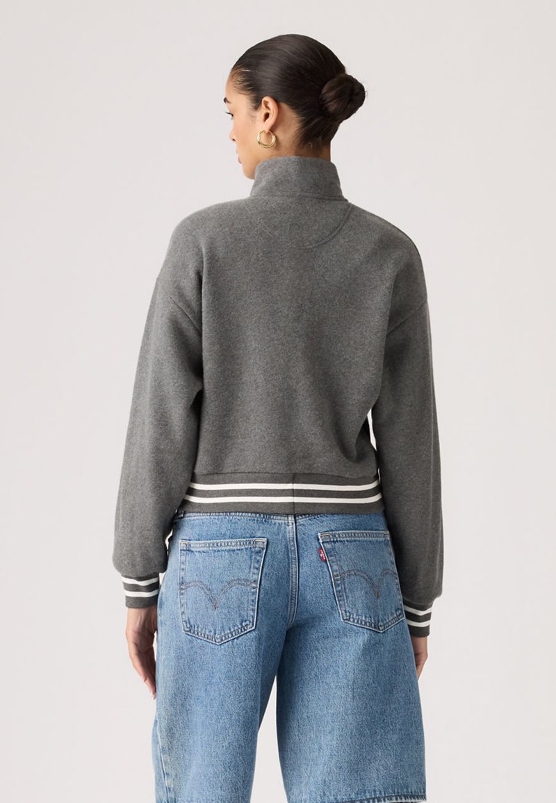 Femme aux cheveux en chignon portant des boucles d'oreilles créoles dorées, un pull gris à col haut avec un ourlet rayé, et un jean en denim bleu clair, vue de dos.