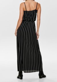 Robe maxi noire à fines rayures blanches verticales, bretelles spaghetti, silhouette fluide et taille cintrée, associée à des bottines à la cheville.