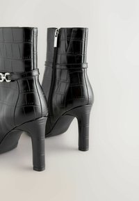 Bottines noires à talons, avec une texture crocodile, fermeture éclair sur le côté et une sangle avec un accent en métal de couleur argent.