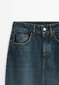 Jean en denim bleu foncé à la taille avec bouton, passants de ceinture, poches avant et coutures contrastantes sur fond blanc.