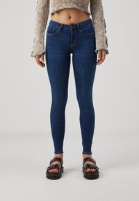 Jeans ajustados de color azul con costuras oscuras, cintura a media altura, dos bolsillos frontales y cierre de botón. Combinados con sandalias marrones con tiras.