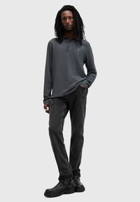 Man met lange dreadlocks, gekleed in een donkergrijze polo met lange mouwen, zwarte jeans en zwarte laarzen, staand met één hand in de zak.