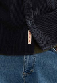 Marineblå kordfløjlsskjorte med en karakteristisk ribbet tekstur, prydet med mørke knapper og et rød-mærket tag, parret med blå denim jeans.