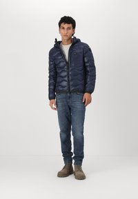 Giacca a piumino blu navy con cappuccio, caratterizzata da un design trapuntato e chiusura con cerniera, abbinata a una camicia di colore chiaro, jeans in denim e stivali marroni.