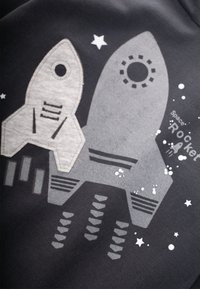 Sudadera gráfica que presenta dos formas de cohetes en tonos de gris, sobre un fondo negro, con acentos de estrellas y el texto que dice "Cohete Espacial."