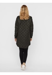 Cappotto lungo imbottito di colore verde scuro con orlo arrotondato. Presenta maniche lunghe e una texture liscia. Abbinato a leggings neri e scarpe leggere.