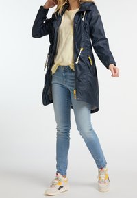 Navy waterproof raincoat with a zipper, drawstring hood, and yellow accents. Styled with light jeans and mixed-color sneakers.