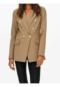 Dwurzędowa marynarka w odcieniu beżowym, z srebrnymi guzikami, klapami z wycięciem oraz bocznymi kieszeniami, zestawiona z czarnymi dopasowanymi spodniami.