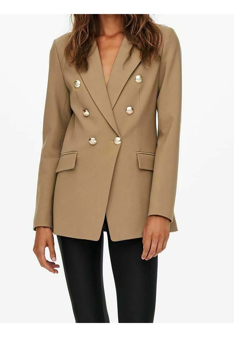 Dwurzędowa marynarka w odcieniu beżowym, z srebrnymi guzikami, klapami z wycięciem oraz bocznymi kieszeniami, zestawiona z czarnymi dopasowanymi spodniami.