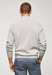 Pull gris clair avec un col côtelé, des manches retroussées et une texture lisse. Porté par-dessus un pantalon bleu avec des poches arrière.