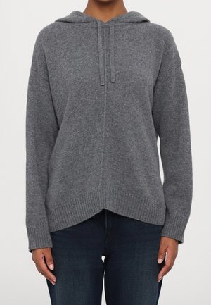Vrouw draagt een grijze gebreide hoodie met trekkoorden en donkerblauwe spijkerbroek, staand tegen een effen witte achtergrond.