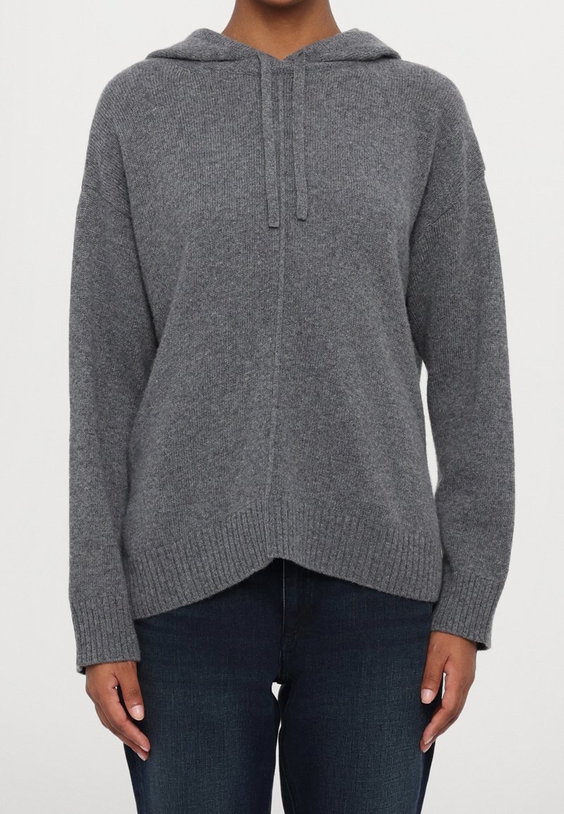 Femme portant un pull à capuche gris en tricot avec des cordons et un jean bleu foncé, debout devant un fond blanc uni.