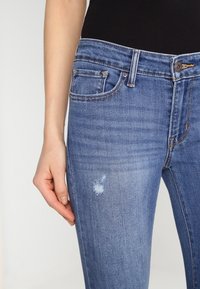 Jeans de mezclilla azul con corte ajustado, que presentan un parche desgastado en el muslo izquierdo, cinco bolsillos y una cintura de altura media.