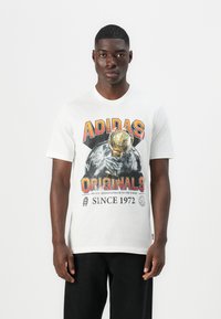 Hvid bomulds T-shirt med grafisk tryk af "ADIDAS" og "ORIGINALS", der viser en hånd, der holder et trofæ. Korte ærmer og rund hals.