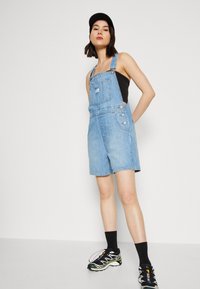 Salopette in denim blu chiaro con vestibilità ampia, spalline regolabili, tasca frontale e bottoni laterali, abbinata a un top nero e sneakers.