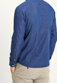 Mężczyzna ubrany w długą niebieską koszulę jeansową i beżowe spodnie, stojący tyłem do widza z rękami w kieszeniach.