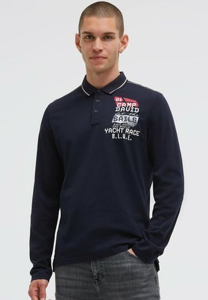 Junger Mann mit kurzen Haaren, der ein marineblaues Polo mit langen Ärmeln und segelbezogenem Schriftzug sowie graue Jeans trägt, steht vor einem einfarbigen Hintergrund.