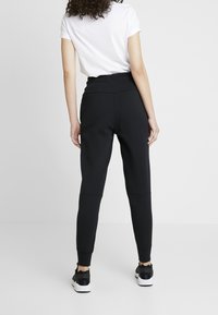 Kvinna som står vänd bort med en vit t-shirt, svarta högmidjade joggingbyxor och svarta och vita sneakers mot en vit bakgrund.