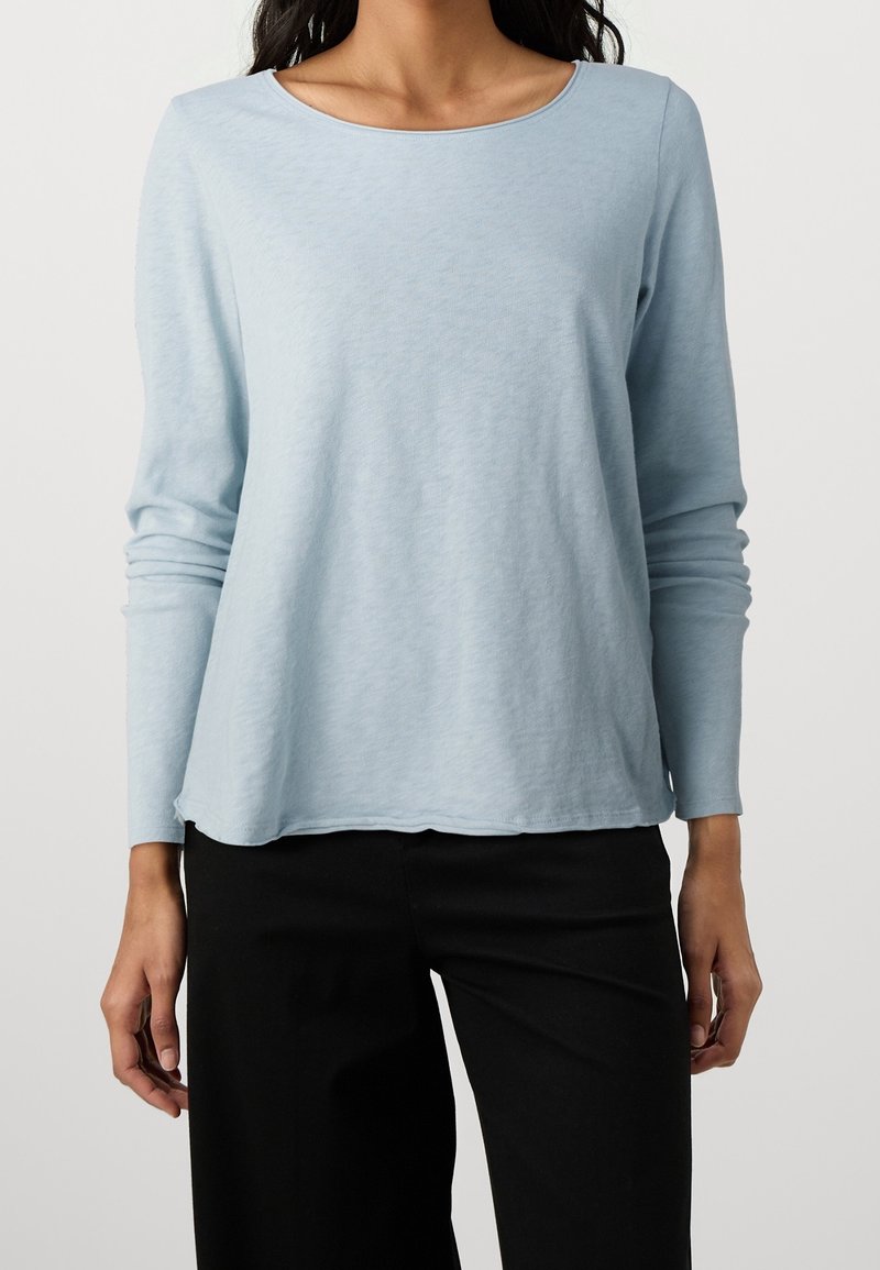 Ljusblå långärmad topp, rund halsringning, mjuk textur, avslappnad passform, svängd fåll, kombinerad med svarta byxor. Inga synliga mönster eller hårdvara.