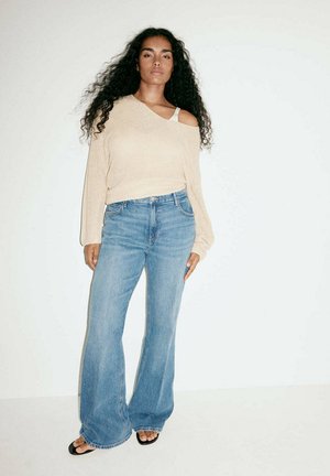 Femme aux cheveux bouclés portant un pull beige épaules dénudées, un jean bleu à jambes larges et des sandales noires, debout sur fond blanc.