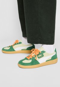 Sneaker in suede verde e crema con lacci arancioni, dettagli bianchi e suola in gomma testurizzata, con logo del marchio sul lato.