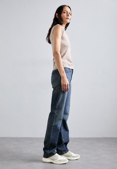 Light beige sleeveless top, blue denim jeans, and light gray sneakers. Model stands in profile against a plain background.