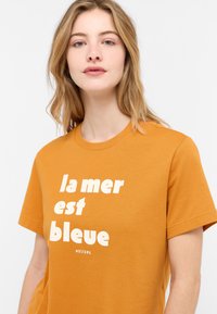Oranžinė Mustang marškinė su trumpomis rankovėmis ir apykakle, kuriuose iš priekio puikuojasi baltas užrašas "la mer est bleue". Pagaminti iš medvilnės, turintys lygią tekstūrą.