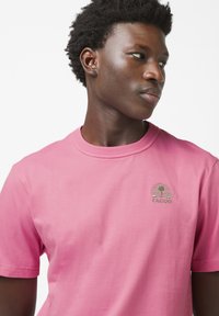 Rosa Baumwoll-T-Shirt mit Rundhalsausschnitt, kurzen Ärmeln und einem grünen gestickten Logo mit einem Baum- und Sonnendesign auf der linken Brust.