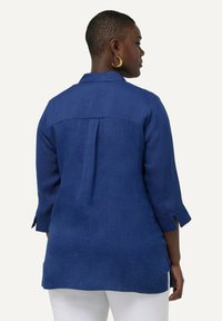 Camisa de lino azul marino con un pliegue en la espalda, mangas tres cuartos y aberturas laterales. Presenta un cuello clásico y una textura suave.