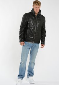 Sort læderjakke med lynlås foran, teksturerede skuldre og en grå hætteliner, parret med lyseblå slidte jeans og hvide sko.