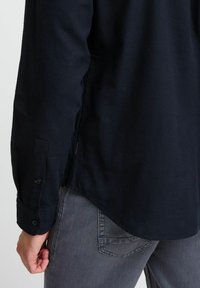 Marineblå bomullskjorte med lange ermer, knappede mansjetter og sideskjitt, sammen med grå skinny jeans med detalj på baklommen.