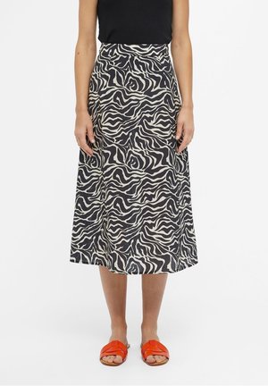 Frau trägt einen schwarz-weißen Zebra-Midi-Rock, ein schwarzes ärmelloses Oberteil und orangefarbene Slip-on-Sandalen, steht vor einem weißen Hintergrund.