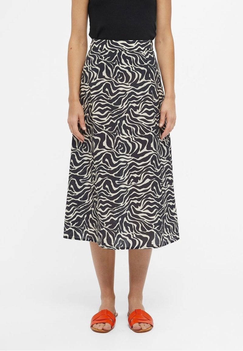 Femme portant une jupe midi à imprimé zèbre noir et blanc, un haut noir sans manches et des sandales orange à enfiler, debout sur un fond blanc.
