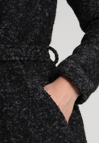 Cappotto nero testurizzato con materiale morbido e peloso, vita aderente, tasca nascosta e collo dal design rilassato.